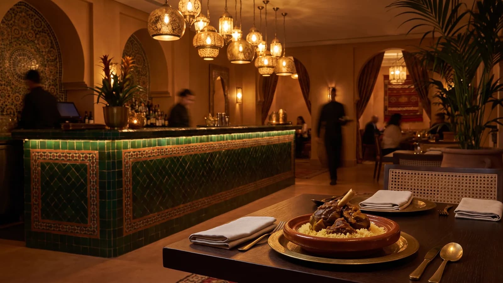 Salle d’un restaurant marocain contemporain en service : bar en zellige, tables dressées, lumière chaude