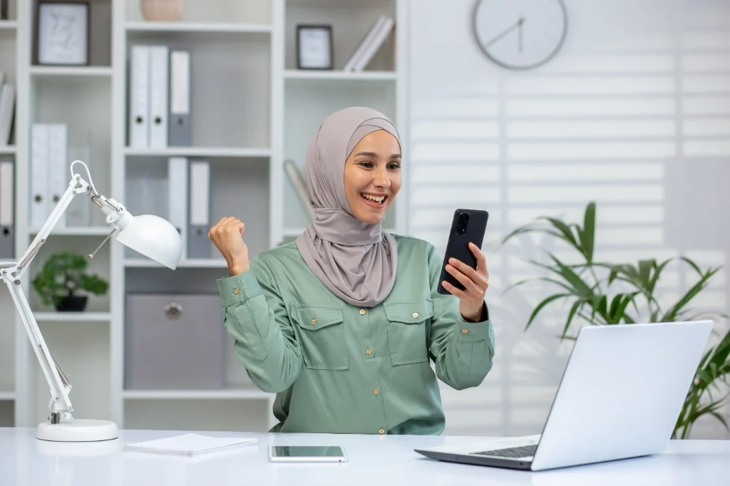 Créatrice professionnelle en hijab, victoire sur smartphone au bureau, laptop et bureautique blanche — contenu de marque et campagne d’influence, cadre professionnel moderne.